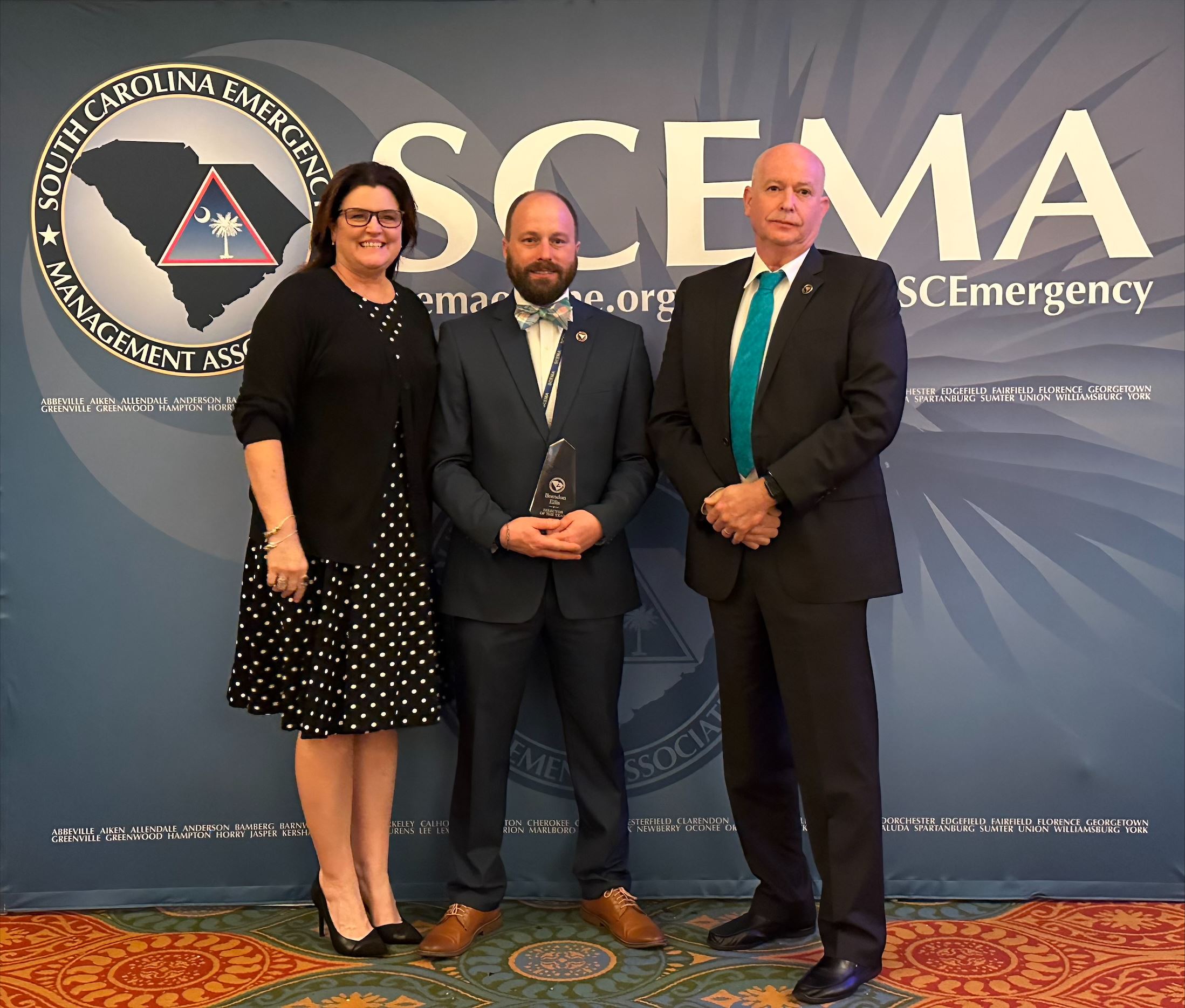 Three people standing in front of a backdrop emblazoned with the SCEMA logo and name