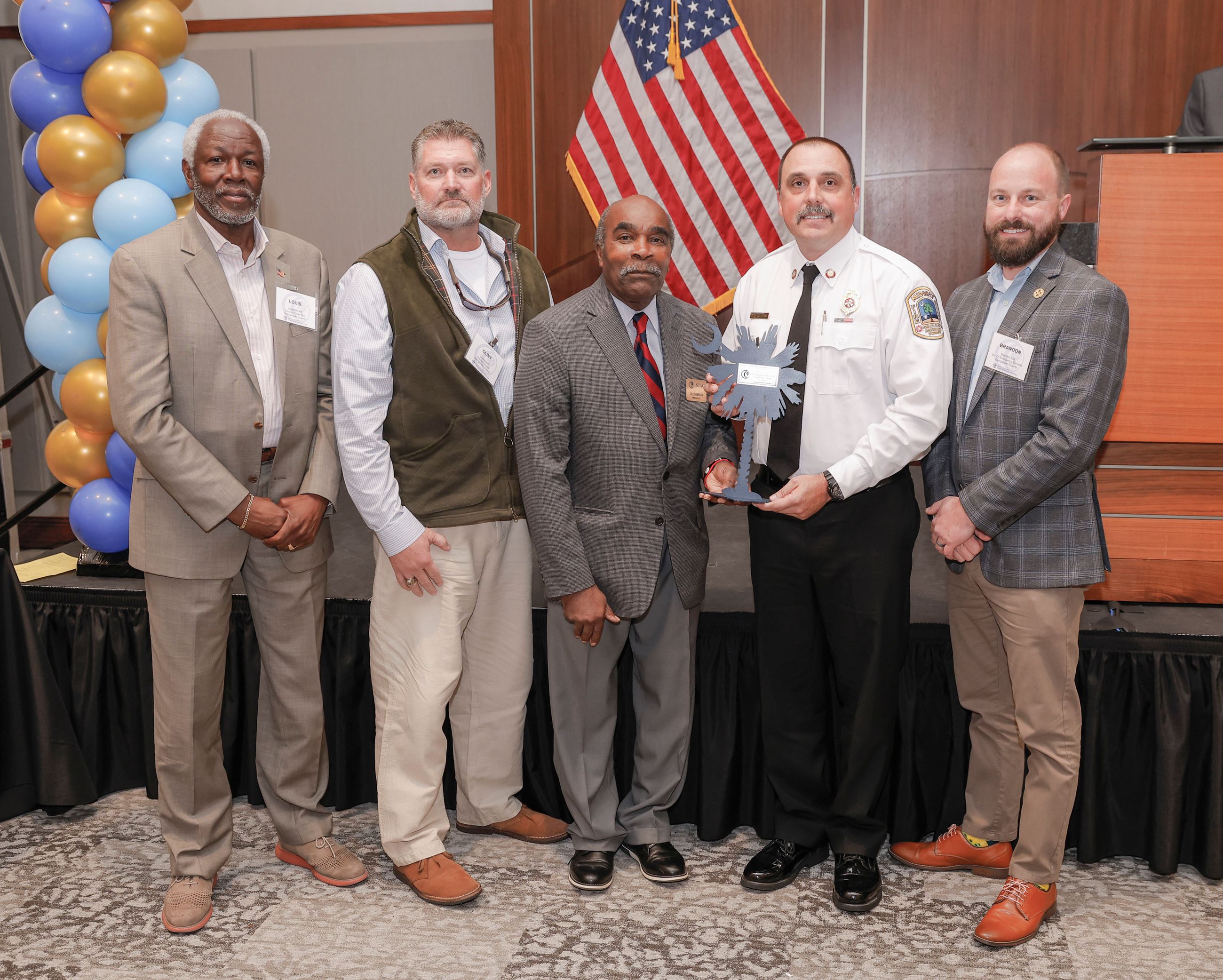 Five men stand in front of an American flag and balloons while one holds an award