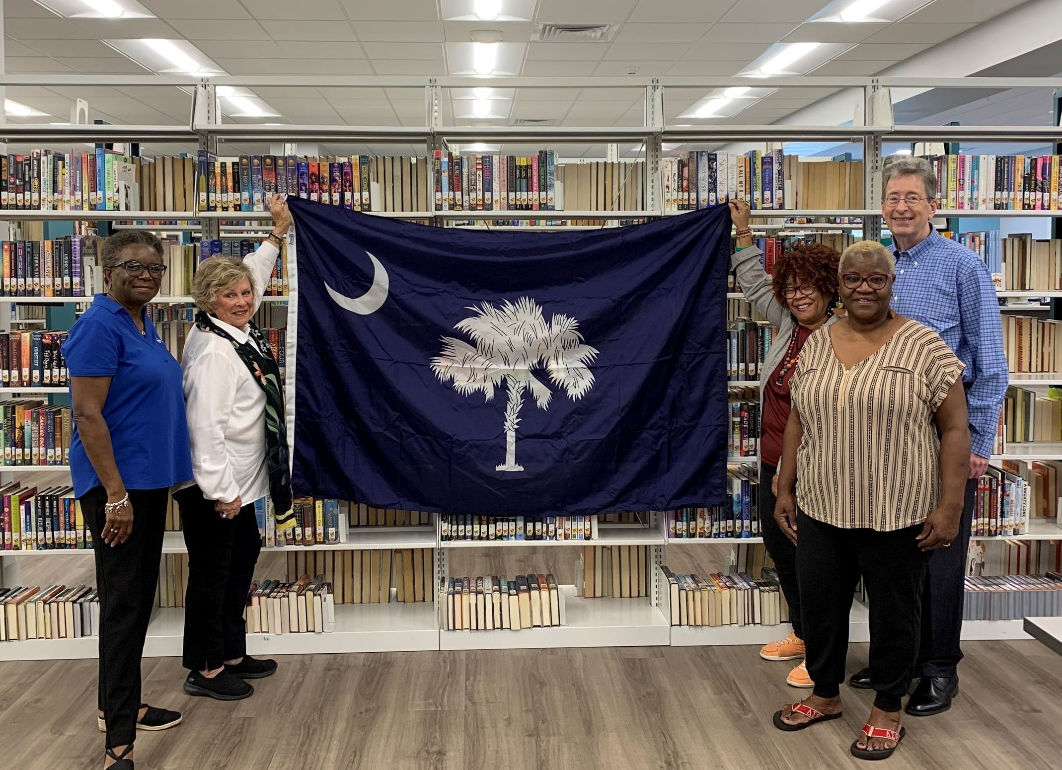 A group of five people holding up a large SC state flag in front of a row of book cases.