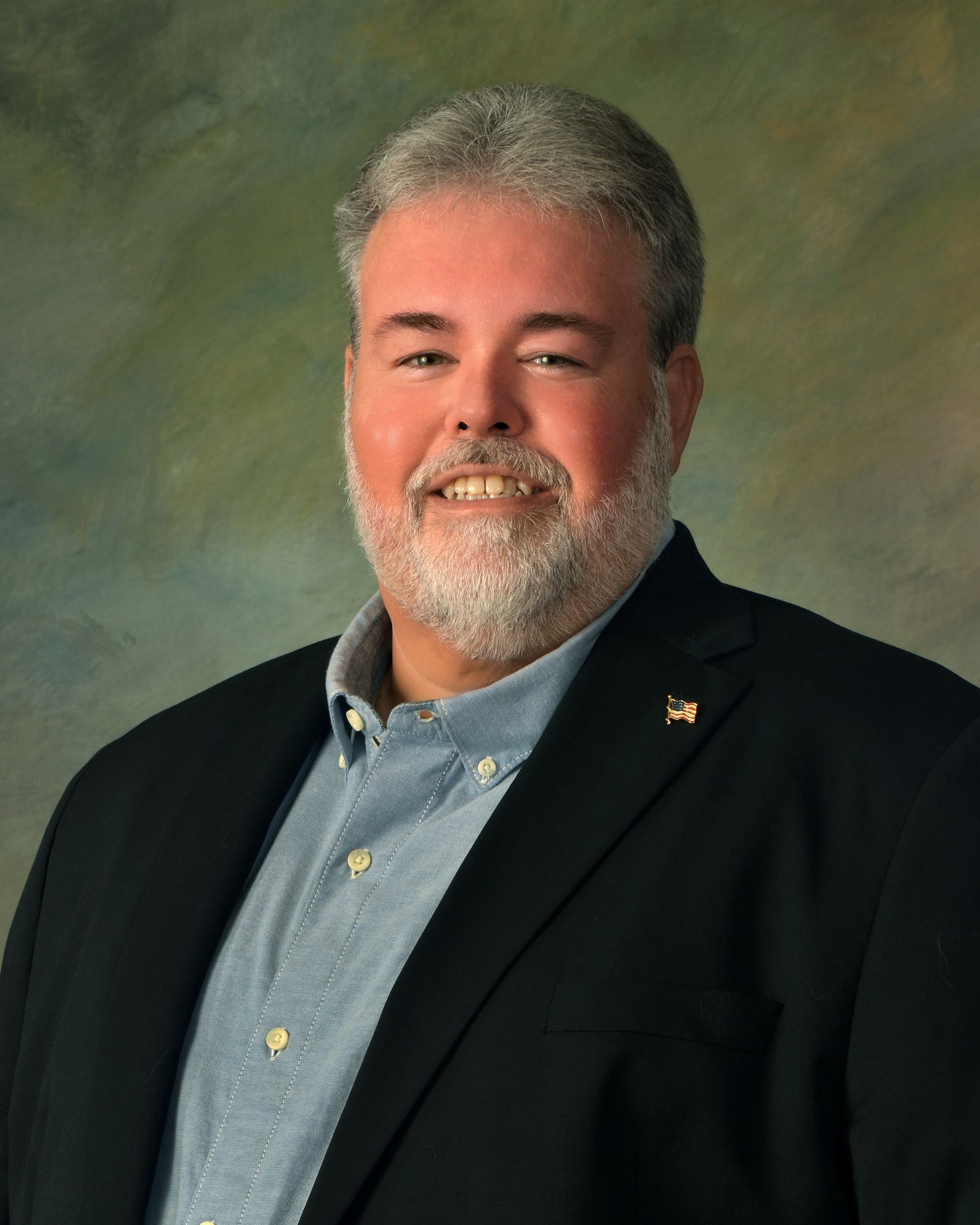 Ron J. Charlton, a white man with grey hair & a grey beard wearing a suit, smiling.